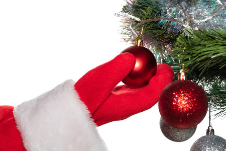 Santa Claus's hand holding a glittering red ball hanging on decorated Christmas tree. Tree, tinsel, and other balls are blurred. Winter holidays decoration concept. Isolated on white background.の写真素材