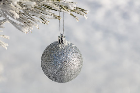 Close up shot of a single white glittering Christmas ball decoration hanging off a Christmas tree outside, partially covered in snow. Tree is also covered with snow. Winter holiday decoration concept.の写真素材
