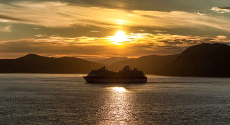 Alaska, USA - June 22, 2018: Celebrity Infinity sailing at sunset in one of the Alaskan Fjords. Beautiful orange sky, sun setting down, and mountains in the background.のeditorial素材
