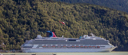 Skagway, Alaska, USA - August 31, 2018: Carnival Splendor docked in the port. Red helicopter flying over her. Massive mountain covered with forest in the backgorund.のeditorial素材