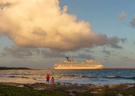 Cozumel, Mexico - February 11, 2020: Carnival Freedom docked in Costa Maya port at orange sunset. Two tourists, shore line, splashing waves in the foreground. Blue cloudy sky in the background.のeditorial素材