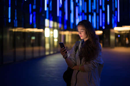 Beautiful young stylish woman walking in the night city streets using modern smartpone, hipster female blogger typing text message on her cellphoneの写真素材