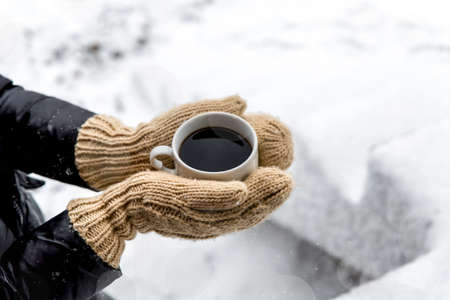 Female hands in woolen knitted gloves holding cup of warm black coffee outdoors on snowy background. Top view of hands and coffee mug. Happy, frosty winter morningの写真素材