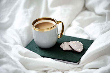Cup of warm coffee, notebook and cookies on white bed linen. Cozy morning at home