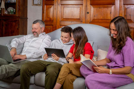 Beautiful family is on the sofa at home - Father is working with laptop, mother is reading a book...