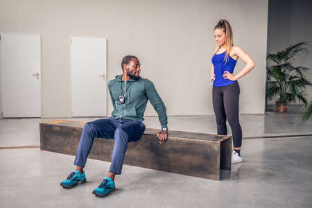 The black coach show to the young girl how to perform the exercise in the gym - Afro american personal trainer is showing the exercise at beautiful girl in the gymの写真素材