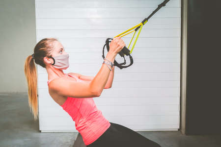 Girl training her arms with protective mask and straps Trx Fitness in the gym during quarantine time for pandemic covid19の写真素材