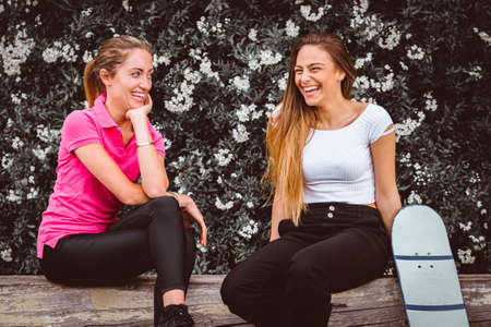 Portrait of two beautiful girls while sitting and talking and joking laughing - Two girlfriends are sitting while talking and laughing with the skateboard in a moment of relaxationの写真素材