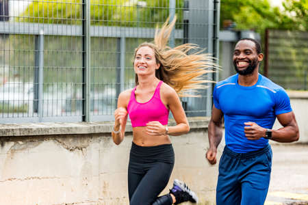 Young couple smiling to each other while jogging on the street, out to run. Afro american man and beautiful blonde girl in sportswear running through the city street together - Health and fitnessの写真素材