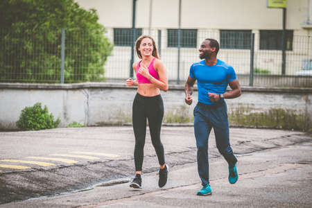 Young couple smiling to each other while jogging on the street, out to run. Afro american man and beautiful blonde girl in sportswear running through the city street together - Health and fitnessの写真素材