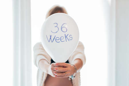 A pregnant woman is holding a balloon with the words 36 weeks - Young pregnant girl is behind and...