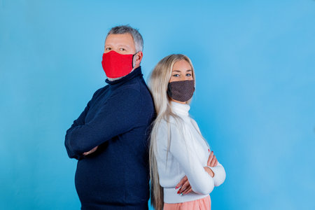 Portrait of hipster man and young girl standing back to back with folded arms wearing colorated face masks isolated on light blu backgroundの写真素材