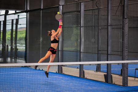 Woman playing padel in a blue grass padel court indoor - Young sporty woman padel player hitting ball with a racketの写真素材