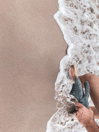 Waves texture on the beach while a man strolls with flip flops in hand - Sandy beach background with white foam wavesの写真素材