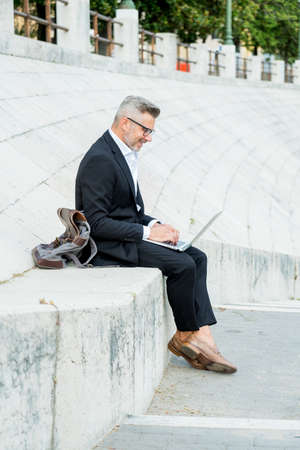 Businessman working laptop computer while sitting on stairs in city centerの写真素材
