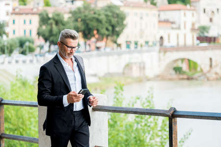 Urban businessman wearing jacket and shoulder bag. Handsome male model in his 45s. Young entrepreneur walking in the street and holding a mobile smartphone using app text sms messageの写真素材