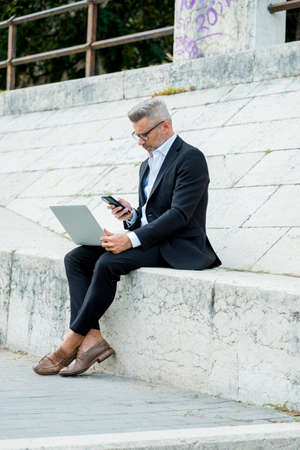 Handsome young businessman sitting on the stairs outdoors working with his laptop computer while checking message on phone. Distance workingの写真素材