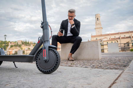 Young businessman sitting on stairs using phone near electric scooter. Focus on scooter