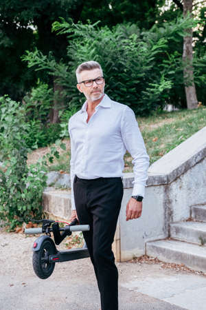Senior businessman holding electric scooter in his hand while going outdoors in the city