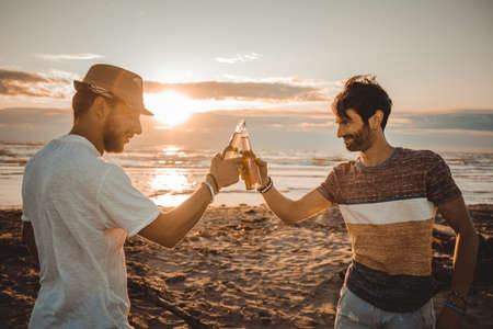 Cheerful young people spending time together on the beach and drinking beer and toasting at the...