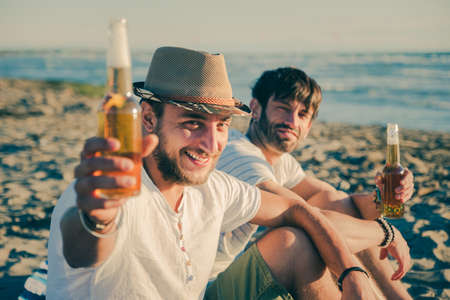 Portrait of friends having a rest on the beach drinking beer. Cheerful young people spending nice time together while sitting on the beach and smiling at cameraの写真素材