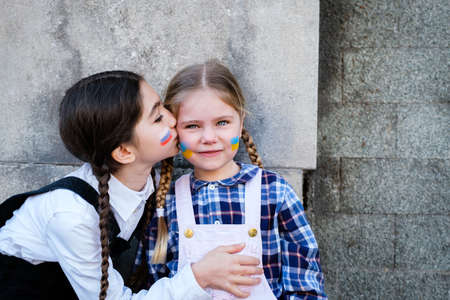 Portrait of two little girls embracing and kissing with Russian and Ukrainian flags on the faces....