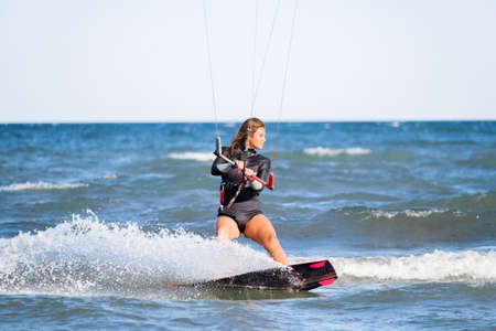Kite surfing girl in swimsuit with kite in the sky rides the waves with splashing water. Water sports, sporty woman, action concept and hard sportの写真素材