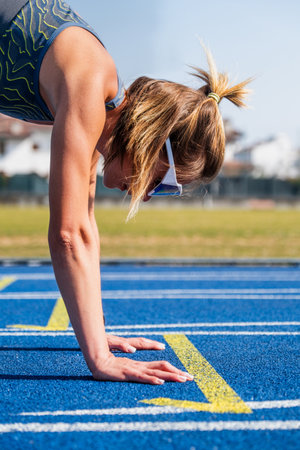 Young athlete woman is doing stretching exercises before starting her race on a blue running track on a sunny dayの写真素材