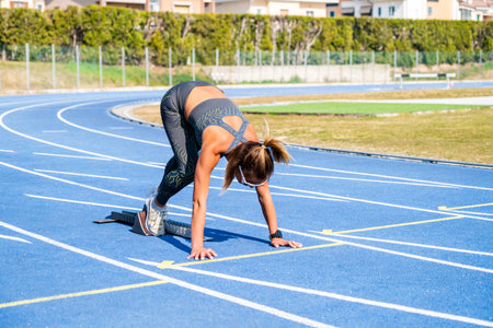 Strong female athlete crouches on a blue track, ready to sprint under the bright sun for a summer...