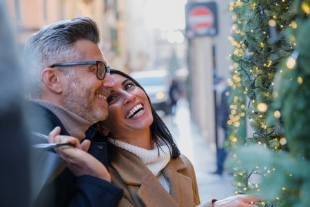 Joyous couple embraces the holiday spirit while exploring the city streets adorned with Christmas lights and decorations, laughing and admiring festive displays as they carry shopping bagsの写真素材