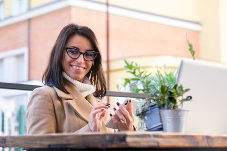 Businesswoman happily multitasks with coffee, laptop, and smartphone at an outdoor cafe, exuding confidence and elegance in a bustling cityの写真素材