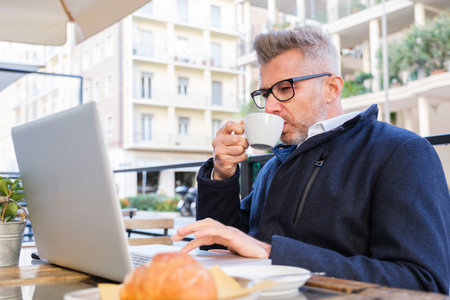 Stylish businessman with gray hair enjoys coffee while working on laptop at outdoor cafe, showing multitasking in urban setting.の写真素材