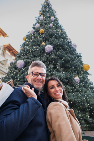 Man and a woman are enjoying christmas shopping. They are posing for a picture in front of a big decorated Christmas treeの写真素材