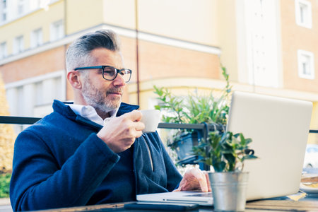 Stylish businessman with gray hair enjoys coffee while working on laptop at outdoor cafe, showing multitasking in urban setting.の写真素材