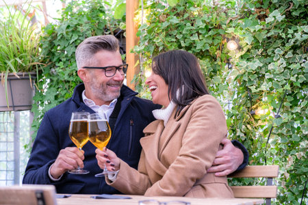 Man and a woman are laughing and clinking glasses of beer together at an outdoor restaurant, surrounded by lush greeneryの写真素材