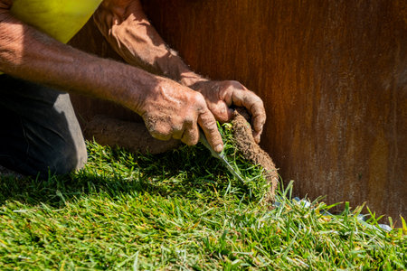 A worker carefully cuts and shapes a piece of turf grass to fit snugly along the edge of a rusted metal planterの写真素材