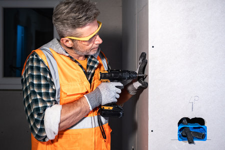 Electrician securing drywall with a cordless power drill. Wearing gloves and safety glasses while working on electrical system installationの写真素材