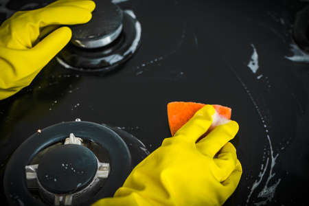 A woman in yellow gloves is cleaning the surface of a gas stove. A sponge and detergent are used. Close-up. Selective focusの写真素材