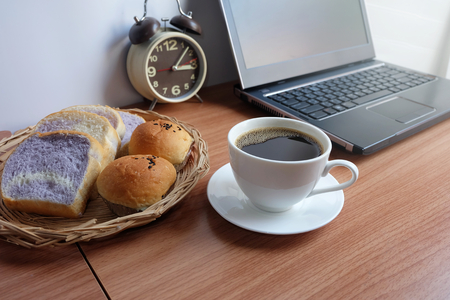 Black coffee in a white cup, bread, and laptop computer on wooden floorの写真素材
