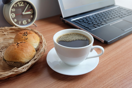 Black coffee in a white cup, bread, and laptop computer on wooden floorの写真素材