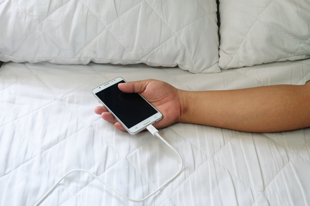 Hand using are holding mobile phones. charger is plugged on the white bedの写真素材