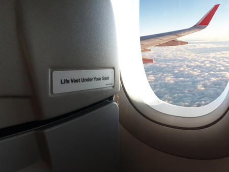 Close up warning words "Life Vest Under Your Seat" On the front passenger seat and Outside the window aircraft wing of the plane with clouds and blue sky and sunlightの写真素材