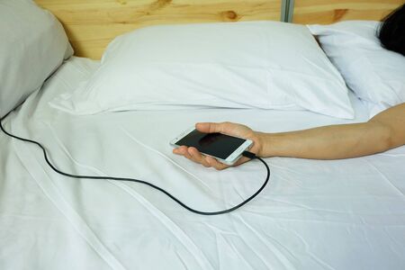 Hand using are holding smart phones. charger is plugged on the white bed, The danger of charging electricity when sleeping.の写真素材
