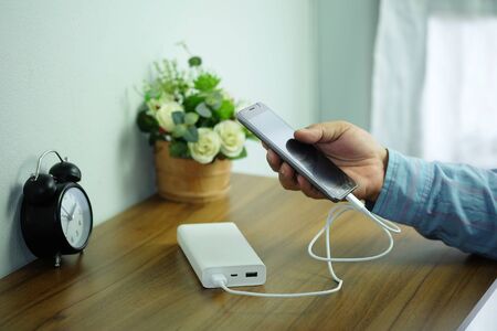 Man is holding smartphone is charging with backup battery or Power bank on the desk wooden table.