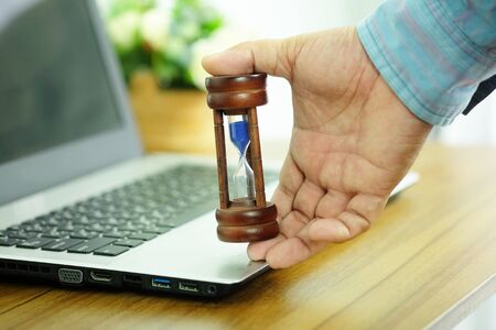 Hand of business man is holding wooden hourglass on laptop computer, Managing time and countdown to...