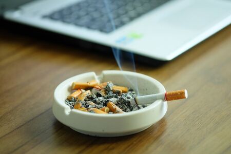 Many cigarette in white ashtray on the desk with laptop computer. Smoking to relieve stress at work but unhealthy.の写真素材