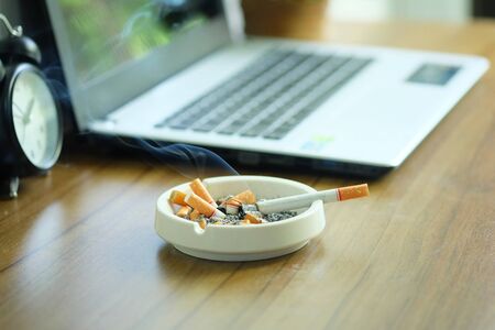 Many cigarette in white ashtray on the desk with laptop computer. Smoking to relieve stress at work but unhealthy.の写真素材