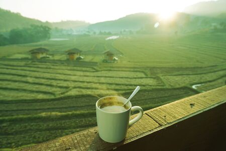 Hot coffee with smoke in white cup on wooden floors and natural rice fields, fresh atmosphere, sunlight in the early morning background.の写真素材