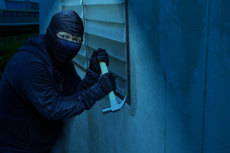 Masked thief are using hammer to break window to steal property in the house at night, detecting CCTV surveillance of home security systemsの写真素材