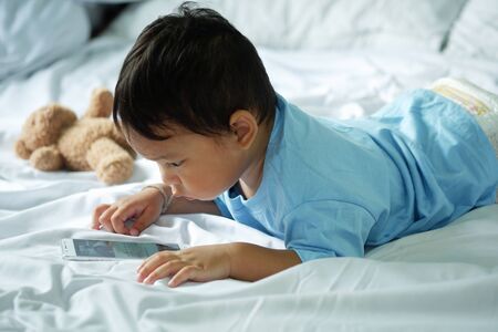 Asian boy about 2 years old, watching and playing mobile phone on the white bedの写真素材